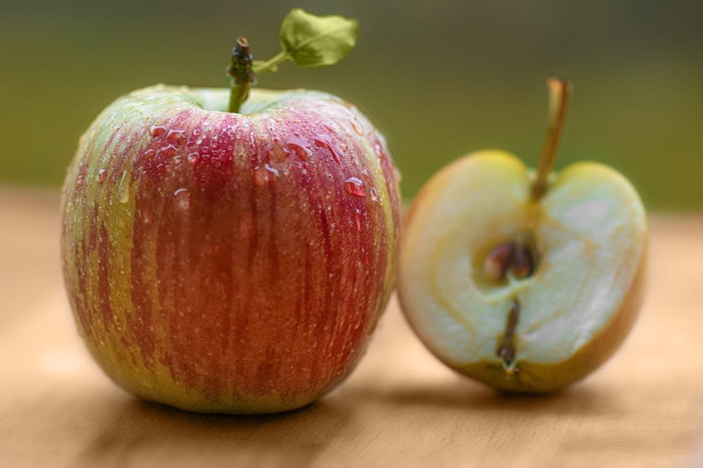 Close-up of a fresh red apple with dew, beside a sliced half on a wooden surface.