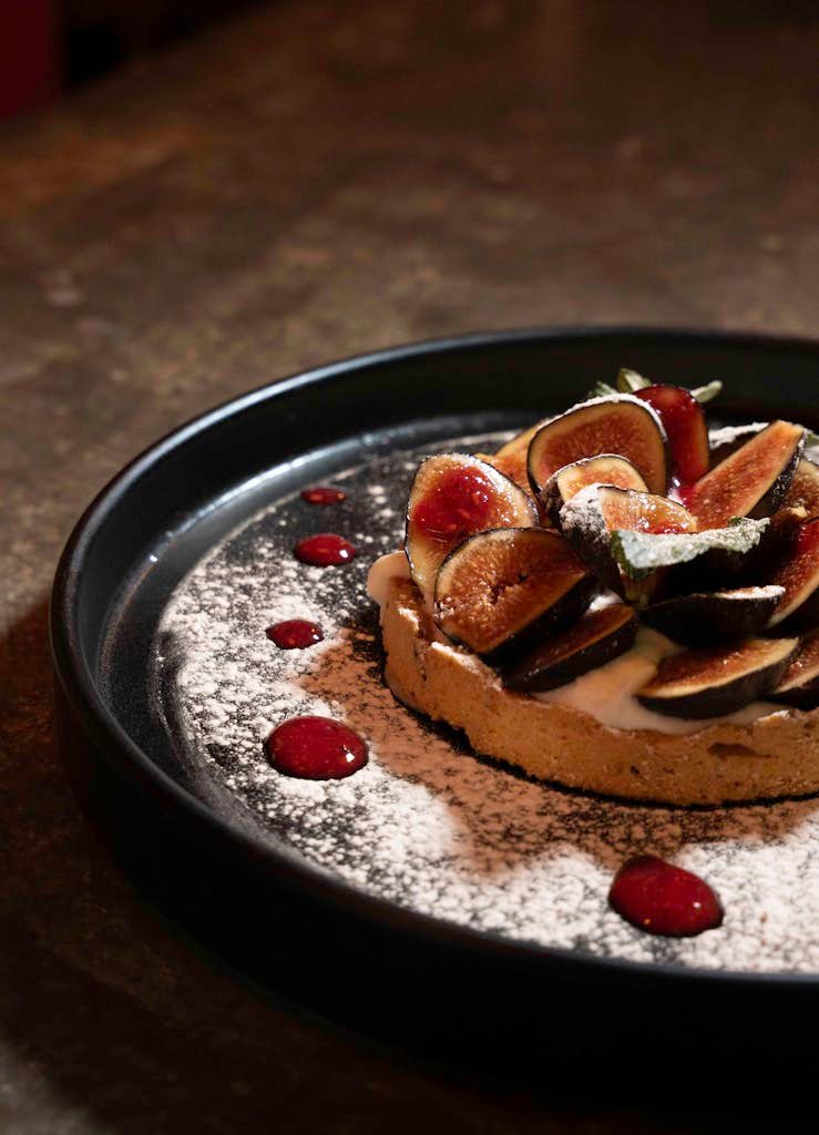 A freshly prepared fig tart with a dusting of sugar and berry coulis on a dark plate.