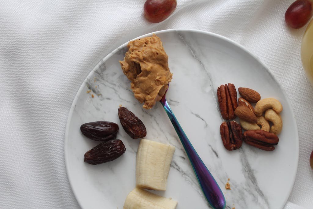 A close-up of a snack plate featuring peanut butter, bananas, dates, nuts, and grapes.