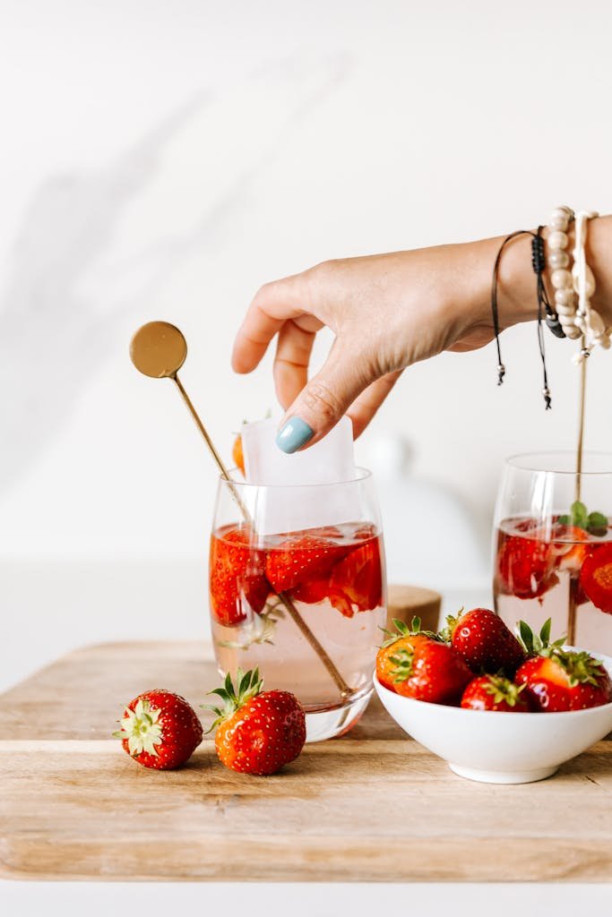 A hand adding ice to strawberry-infused water in a glass, perfect for a refreshing summer drink.