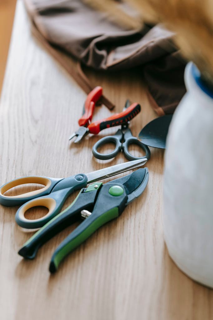 High angle of various gardening tools including scissors wire cutters and pruner placed on wooden table with vase of dry branches