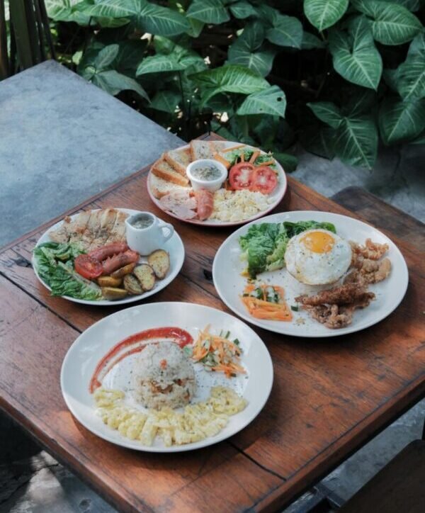 Four plates of Asian breakfast with rice, eggs, vegetables, and meat on a wooden table outdoors.